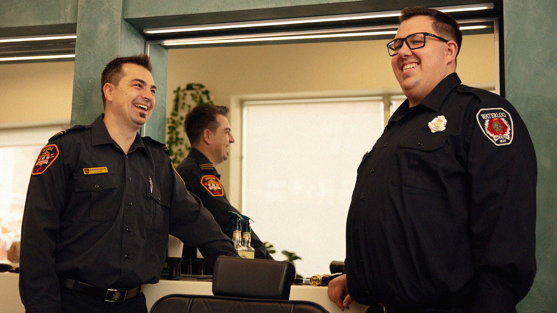 Two firefighters chatting and laughing, standing in front of a mirror at a barbershop